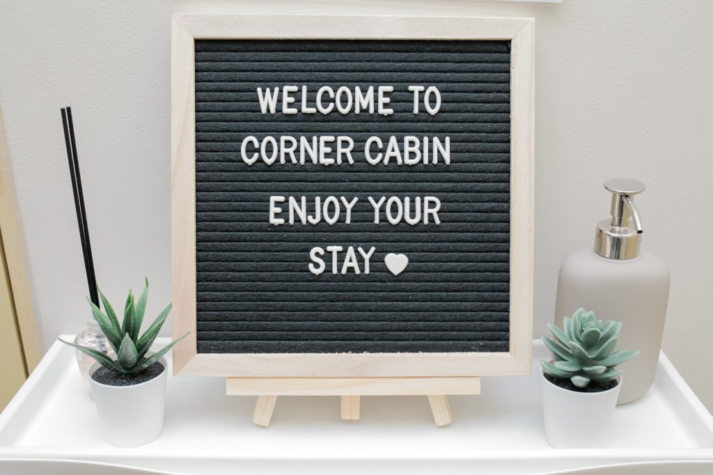 A cozy corner cabin interior featuring a black letter board with a welcoming message that reads "WELCOME TO CORNER CABIN ENJOY YOUR STAY".